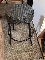Front and side view of the metal and wicker stool standing upright on carpet. Shows round woven wicker seat and black metal legs.