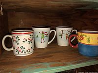 Photo of four decorative ceramic coffee mugs on a wooden shelf, each mug has a different colorful design.