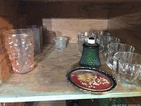 Top view of assorted glassware and decorative items including tumblers, votive holders, green hobnail glass container, and decorative plate on wooden shelf.