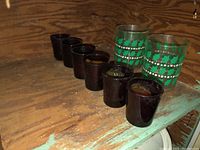 Seven dark brown glass cups lined up next to two green patterned glass tumblers on a wooden shelf.