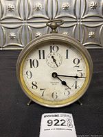 Front view of antique Westclox mechanical alarm clock showing white dial, black numerals, and hands, against a decorative metallic background.