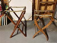 Pair of antique wooden luggage racks, one with turned wooden legs and woven strap, the other with simpler wooden frame and fabric strap, placed on cement floor with some background items.