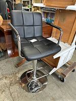 Black adjustable beauty chair shown from front, featuring square tufted padded synthetic leather seat and back, padded armrests, and chrome base.