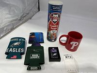 Lot items including the Eagles and Bud Light koozies, Phillies Cooling Towel container, red Phillies mug, Zippo lighter in packaging, and lanyard.