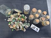 View of nine small glass jars with cork stoppers, two larger jars (one with hinged lid), sea glass pieces and small stones on black cloth.