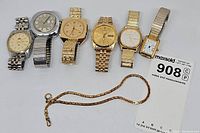 Full view showing six wristwatches and a gold-tone pocket watch chain arranged in a row on white surface with a lot number card.