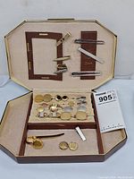 Open jewelry case showing interior compartments with multiple men's tie clips and cufflinks in various gold and silver tones.