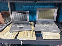 Image of two vintage Apple laptops on a shelf along with two cream-colored Apple external keyboards and an external CD-ROM drive. The laptops appear aged, with a matte gray finish and built-in keyboards and trackpad/trackball. The keyboards and CD-ROM drive show yellowing associated with age and use.