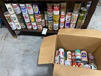 Vintage beer cans lined up on a shelf with additional cans in a cardboard box in front of it