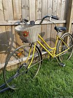 Left side angle view showing the yellow Schwinn Collegiate bike outdoors. The front wicker basket with flower decoration is visible, along with the front wheel and black saddle.