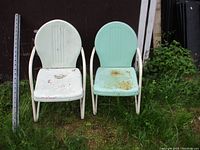 Two vintage metal chairs placed outdoors on grass in front of a dark wall, showing white and turquoise paint with rust and chipping.