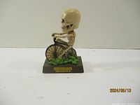 Front and side view of skeleton bobble head riding bicycle on green foliage base labeled 'BONESHAKER'.