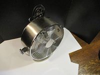 Side angle view of the chrome electric fan showing metal grill, blades and base with power cord in background.