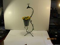 Front view of the metal garden ornament shaped like a faucet holding a flower pot with artificial yellow flowers, standing on a tripod base.