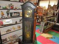Full view of the grandfather clock in a cluttered room environment showing its full height and details of the case, weights, and pendulum behind glass.