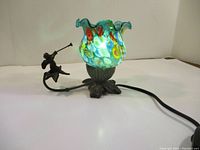 Side view of lamp showing blue-green glass shade and attached cherub figure blowing a trumpet, with power cord visible.