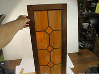 Hand holding the stained glass panel upright showing its wooden frame and pattern of swirled amber glass with three diamonds.