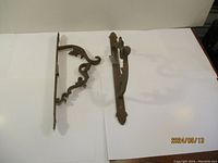 Two metal wall hooks with scroll ornamental metalwork, side views showing mount and hook structure