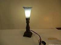 Front view of the tulip-shaped lamp lit up, showing its glass shade and ornate base with power cord.