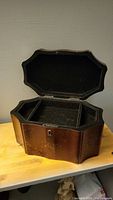 Photo showing open wooden jewelry box with curved octagonal shape, black fabric lining inside compartments, and lock but no key.