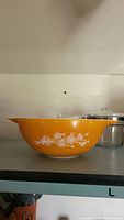 Side view of large bright orange Pyrex mixing bowl with white floral design and wide pouring spout on one side. Orange exterior and white interior.