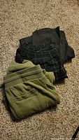 Three pairs of men's pants folded on carpet: one olive green fleece with Berch brand tag, one black with white grid pattern, one black with white pinstripe pattern.