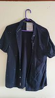 Front view of black short sleeve shirt hanging on purple hanger showing button-up style and collar.