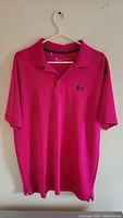 Bright pink short sleeve polo shirt on hanger, front view with collar and Under Armour logo.
