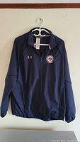 Front view of navy blue Under Armour jacket hanging on white hanger against wall, showing logo on chest and zipper.