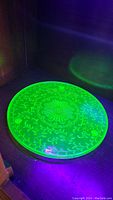 Top view of large circular uranium glass tray glowing bright green with etched floral patterns under UV light.