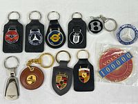 Full lot of 10 automotive keychains and 2 emblems on a white surface with visible brand logos and details.