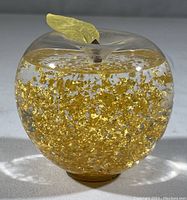 Front view of transparent apple shaped glass snow globe with suspended gold leaf flakes, showing applied gold leaf and stem on top.