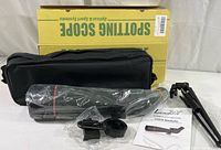 Full lot showing sealed spotting scope in plastic, small tripod, carrying pouch, and manual with yellow original box in background.