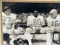 Black and white photo showing three 49ers players sitting on a bench on the sidelines, close-up of their upper bodies and helmets below.