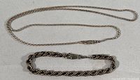 Full set of a slender sterling silver rope chain necklace next to a thicker sterling silver rope chain bracelet both with lobster claw clasps laid flat on a white surface.