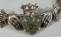 Close-up image showing the Claddagh design with a green stone heart held by two hands and topped with a crowned detail. The silver is stamped.