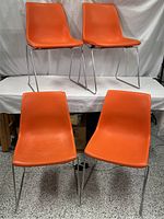 Four vintage orange molded plastic Mid Century Modern shell chairs with chrome wire sled bases arranged in two rows on white fabric backdrop.