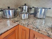 Four stainless steel pots shown: three stock pots varying in size with two lids, and one smaller saucepan with lid. Black handles and knobs.