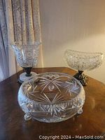 Three cut crystal serving dishes shown on a wooden surface. One round dish with starburst cut pattern and two elevated bowls with decorative metal and marble bases.