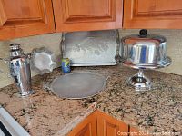 Image showing all items: silver plated jug, small scalloped dish, round tray, large decorated rectangular tray, and pedestal cake plate with dome.