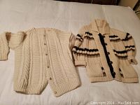 Two handknit women's cardigans laid flat, showing cream Aran-style cable knit sweater and cream sweater with brown stripes.