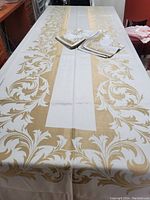 Full view of the rectangular damask tablecloth on a dining table with matching napkins placed on top, showing white fabric with metallic gold foliate pattern along edges.