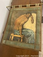 Full view of the fabric wall hanging showing seated male figure in blue pants leaning on a white cushion with decorative borders and hanging rod.