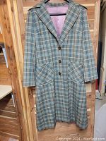 Front view of the full plaid wool tweed trench coat hanging on a wooden hanger. Shows front buttons, patch pockets, and collar details.