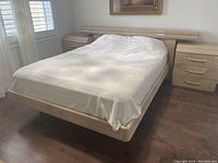 Photo of a light-colored veneer bed frame with a sculptural headboard and mattress covered with white bedding in a bedroom setting.