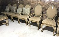 Six wood chairs in a basement with plastic-covered walls. Four side chairs and two arm chairs with carved legs and beige patterned upholstery.
