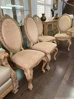Four dining chairs with carved wood frames and beige patterned upholstery. Oval backs and carved legs with claw feet shown, highlighting condition and detail.
