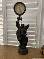 Full view of battery operated pendulum clock mounted on winged couple figurine, showing clock face and pendulum.