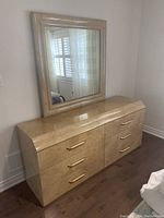 Light-colored veneer dresser with six drawers and gold handles shown at an angle with mirror on top.