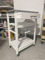 Side view of white kitchen cart showing drawer, wire basket, slatted shelf, and caster wheels in a garage setting.